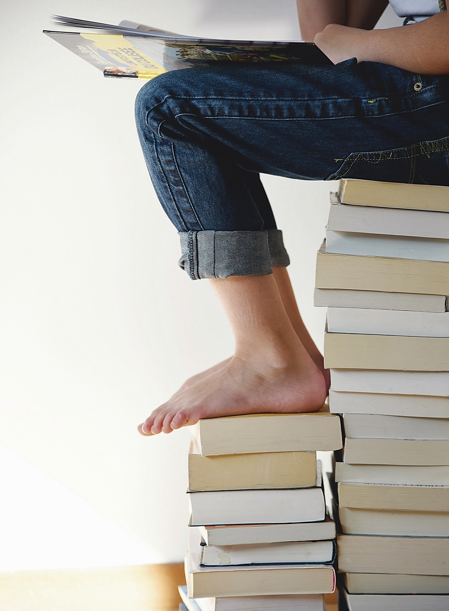 Child sitting on a pile of books reading a book, shown from waist down