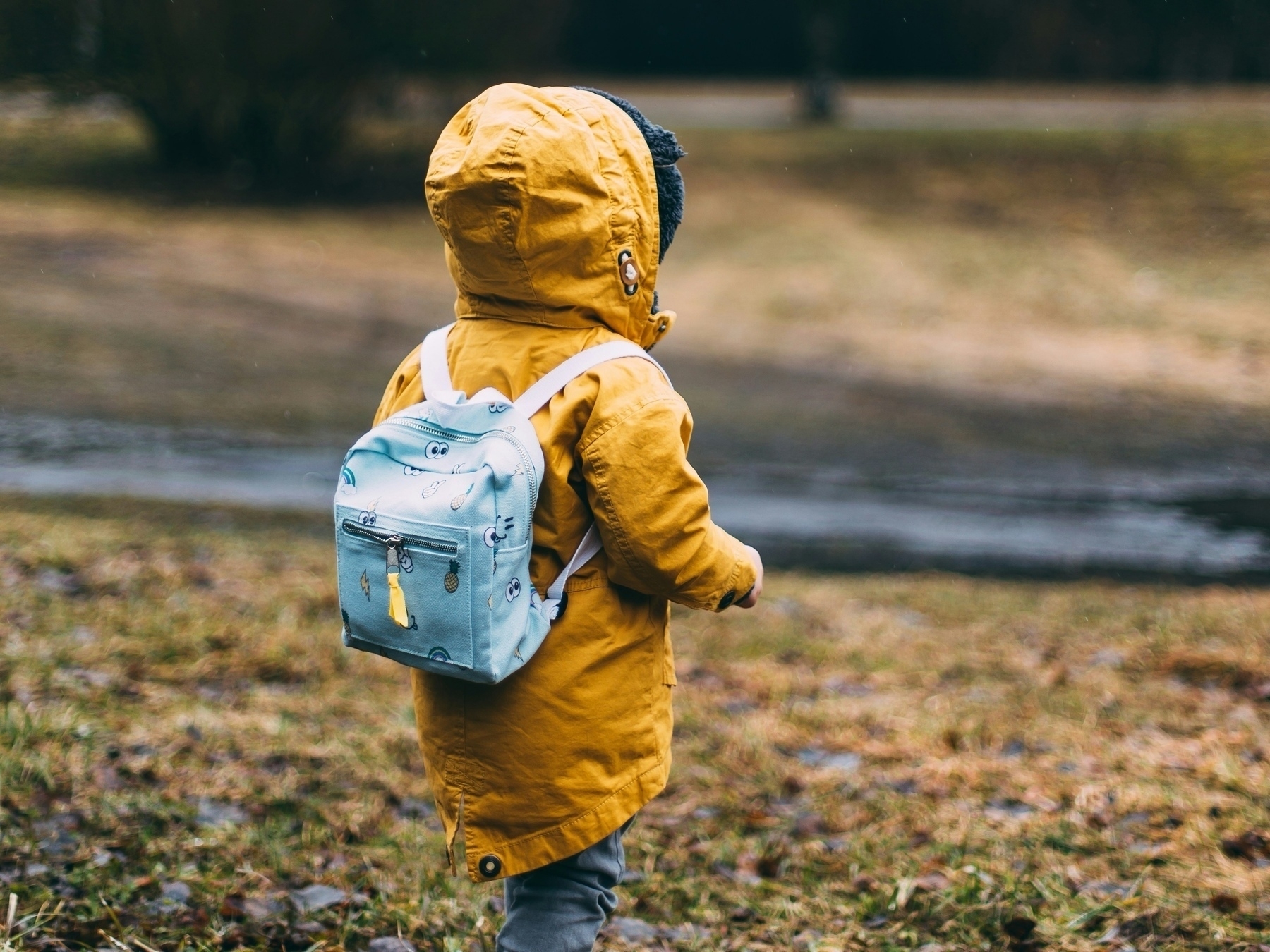 Small child wearing a colorful backpack and facing away from the viewer in an outdoor setting